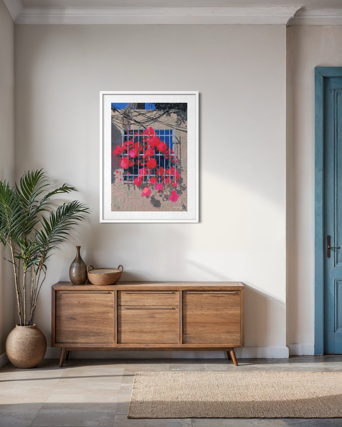 Framed artwork on a wall above a wooden sideboard in a room with a plant and blue door.