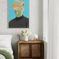 Framed artwork of a person with colorful flowers replacing their head above a wooden side table with a vase of flowers.