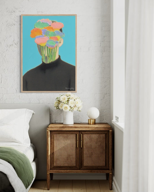 Framed artwork of a person with colorful flowers replacing their head above a wooden side table with a vase of flowers.