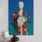 Colorful abstract painting of a figure holding a clock on a wall above books and vases.