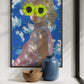 Framed artwork of a person with floral sunglasses on a wall above a shelf with a vase and bowl.