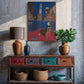 Decorative console table with vases, a lamp, and a painting in a room with concrete walls.