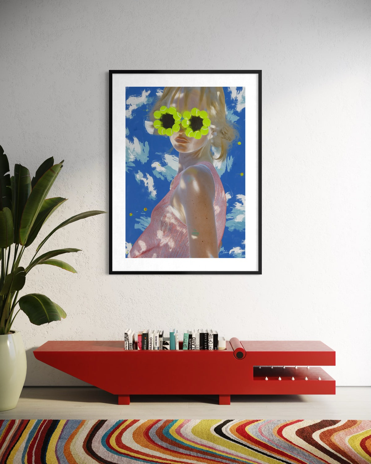 Framed artwork of a person wearing sunglasses with a blue sky background, placed on a red console table.
