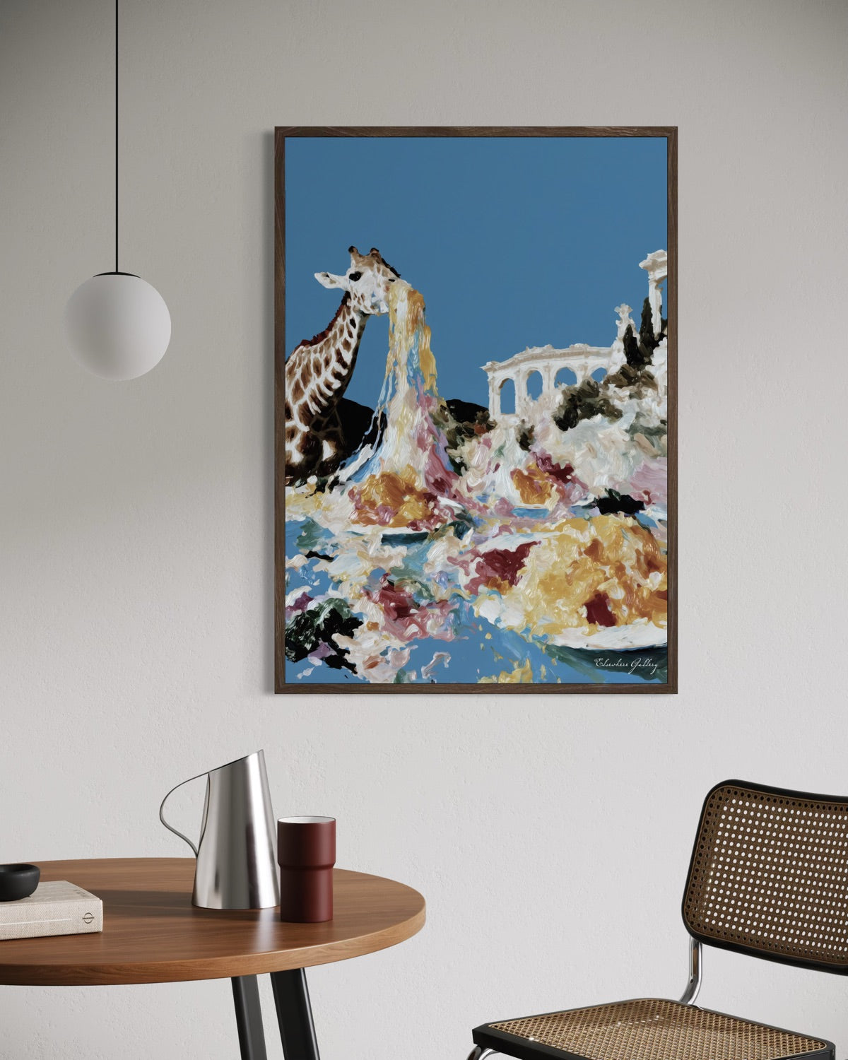Framed artwork of a giraffe in a scenic setting on a wall above a table with a chair.