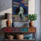 Decorative console table with a colorful painting of a dog and fish, vases, and plants against a textured wall.